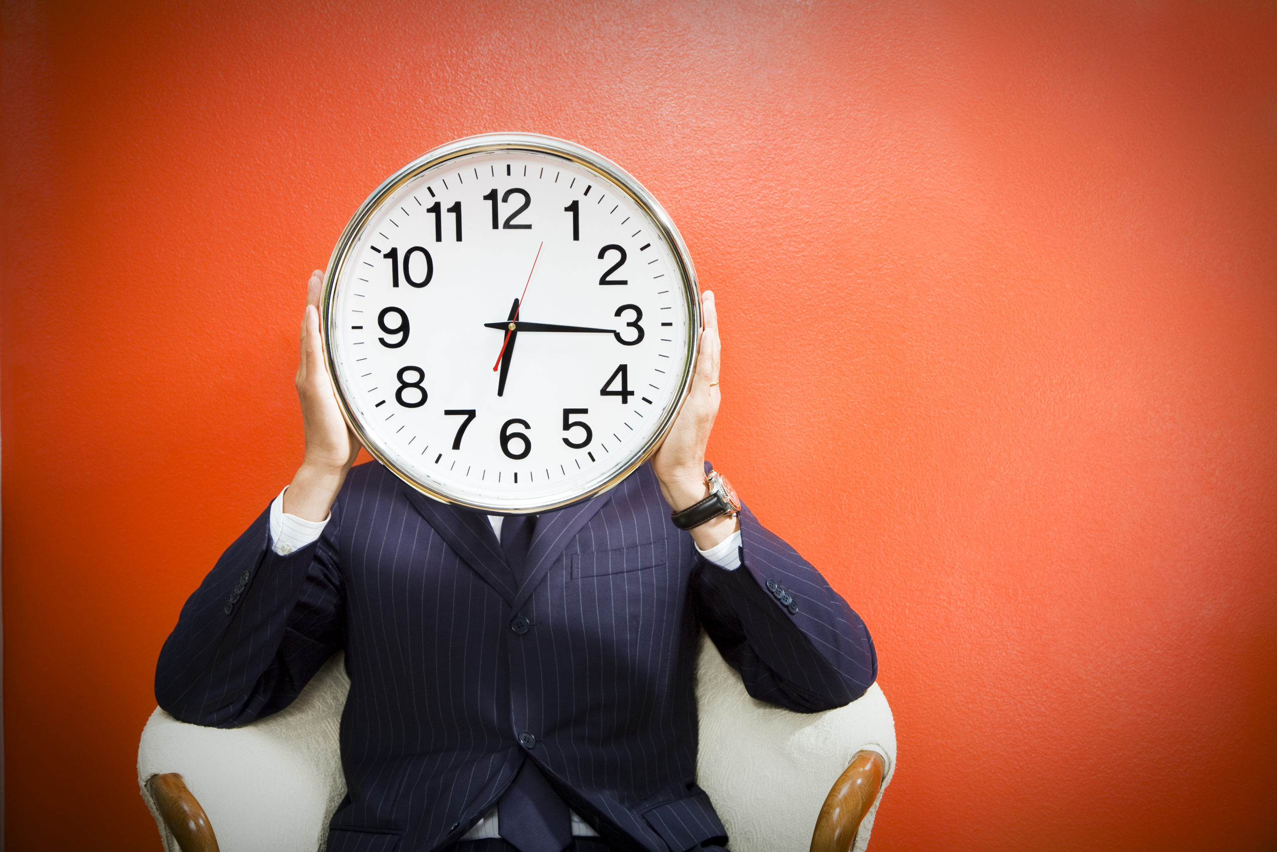 A man in a blue suit holding a clock in front of his face