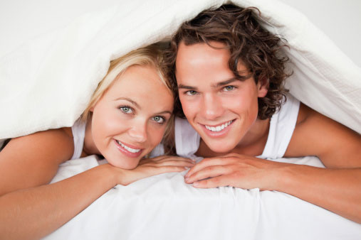 Man and woman laying under some white bed sheets, smiling at the camera